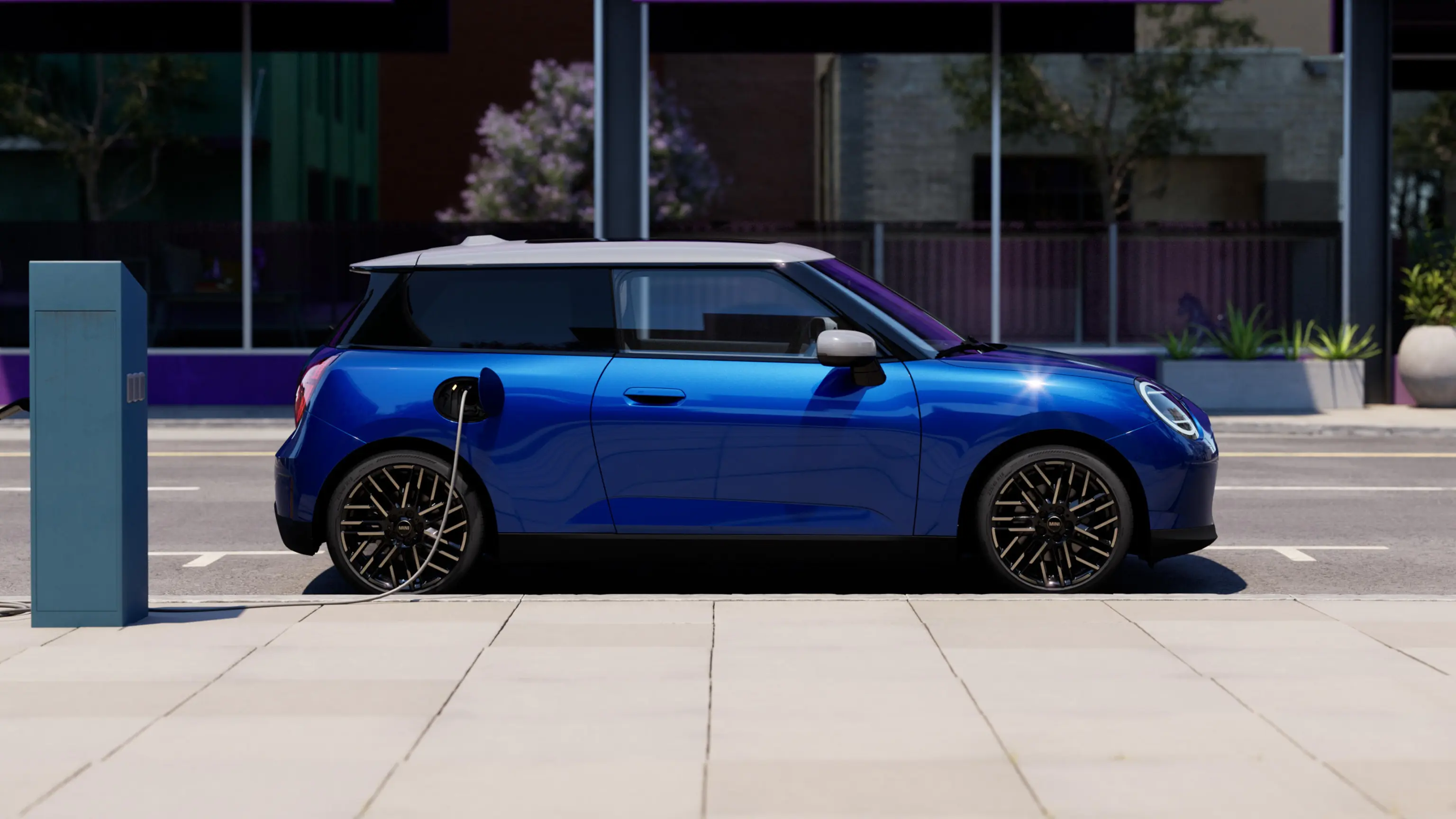 Bild från sidan på helt elektriska MINI Cooper i Blazing Blue parkerad på trottoaren bredvid en laddstation