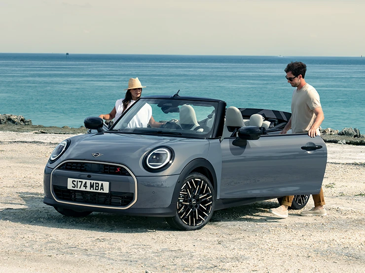 Vy framifrån vänster av en MINI Cooper Cabrio i Copper Grey på en strand med två personer bredvid.