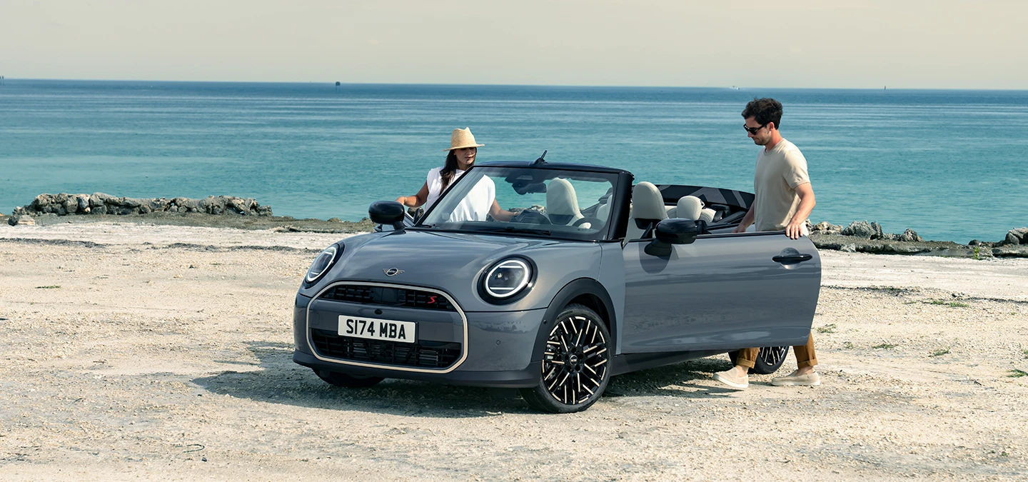 Vy framifrån vänster av en MINI Cooper Cabrio i Copper Grey på en strand med två personer bredvid.
