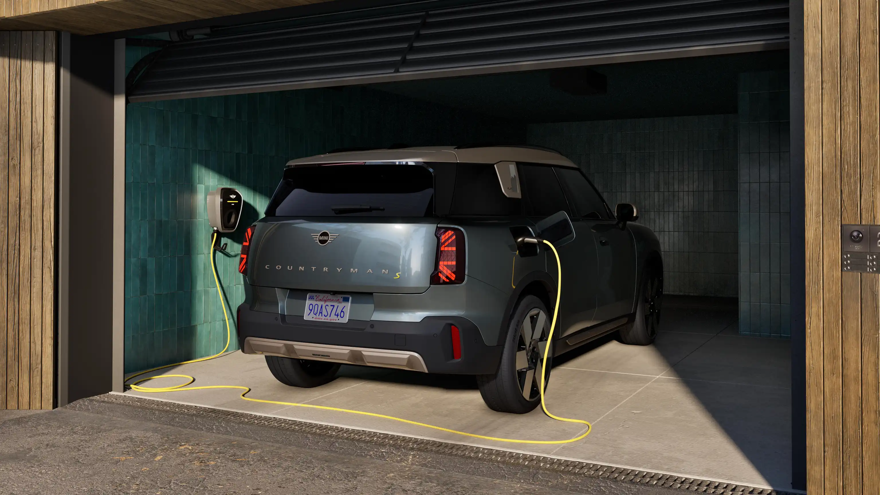 Vy bakifrån av en helt elektrisk MINI Countryman i Smokey Green som står i ett öppet garage och laddar med en ansluten laddkabel.