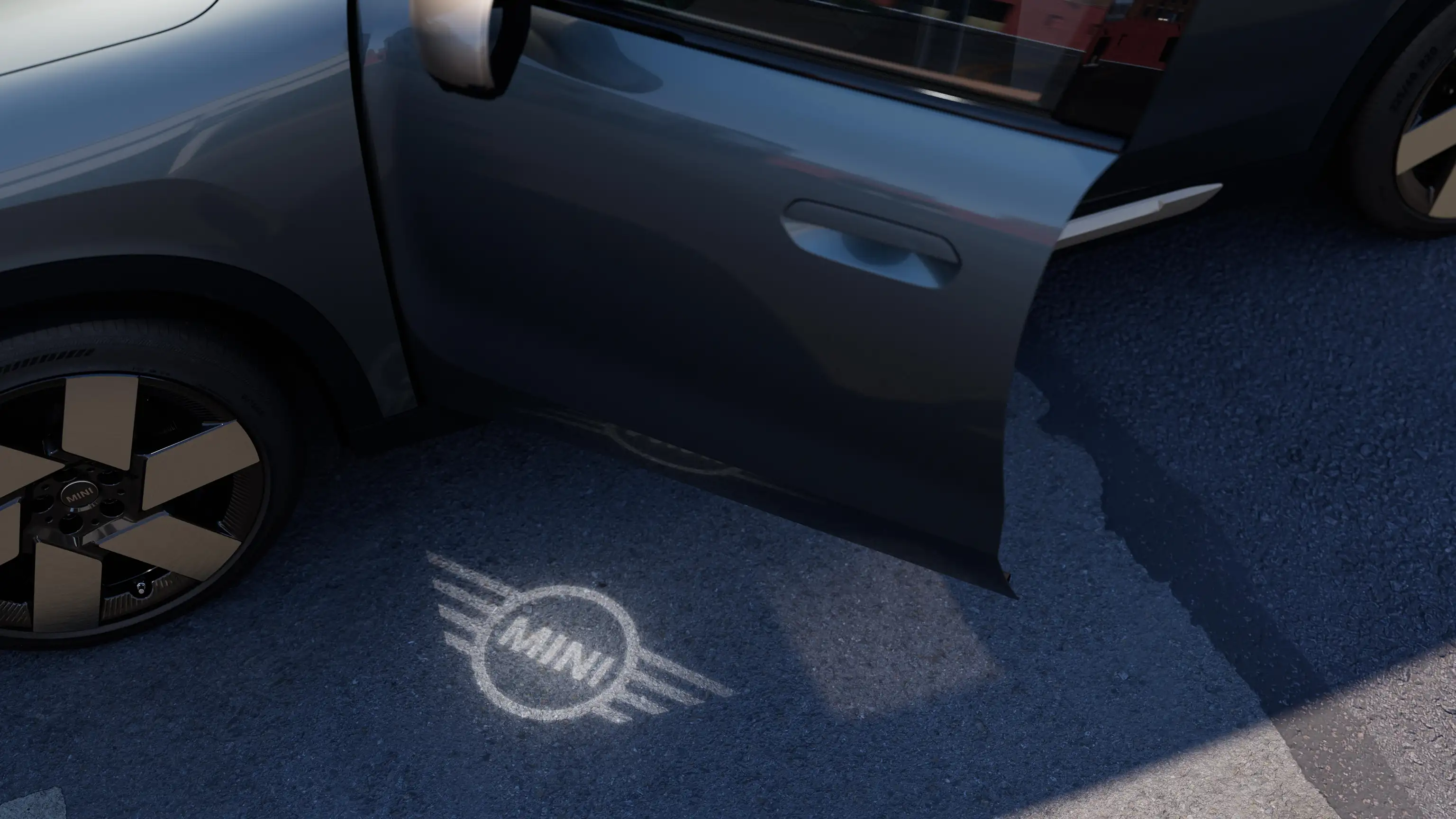 Exteriörvy över helt elektriska MINI Countryman med öppen dörr och välkomstbelysning i form av MINIs logotyp på marken
