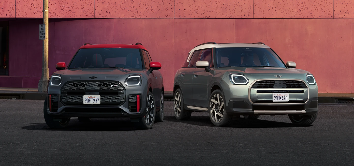Vy framifrån av en MINI Countryman JCW i Legend Grey och rött tak och en MINI Countryman i Smokey Green och tak i Vibrant Silver som står bredvid varandra.