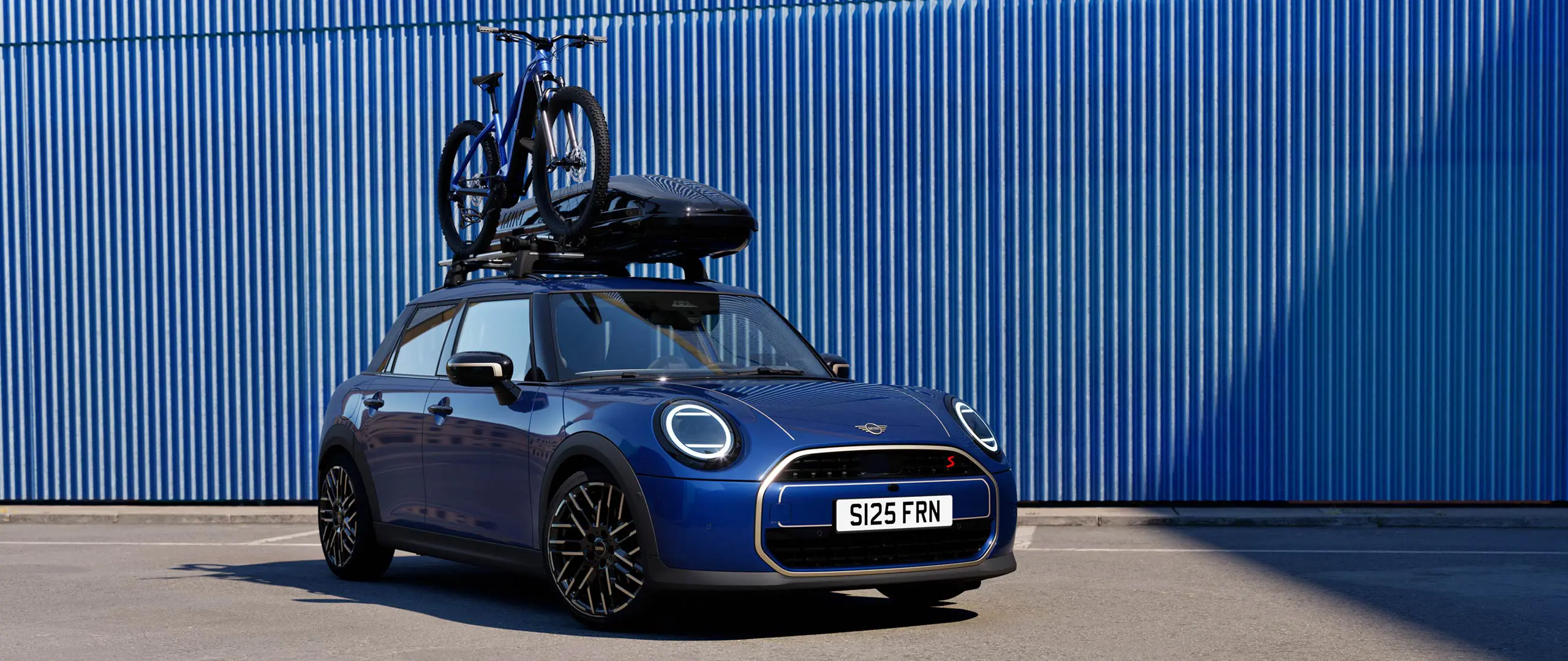 Frontvy av MINI Aceman i Indigo Sunset Blue med MINI takcykelhållare, med en mountainbike, och MINI takbox 320.