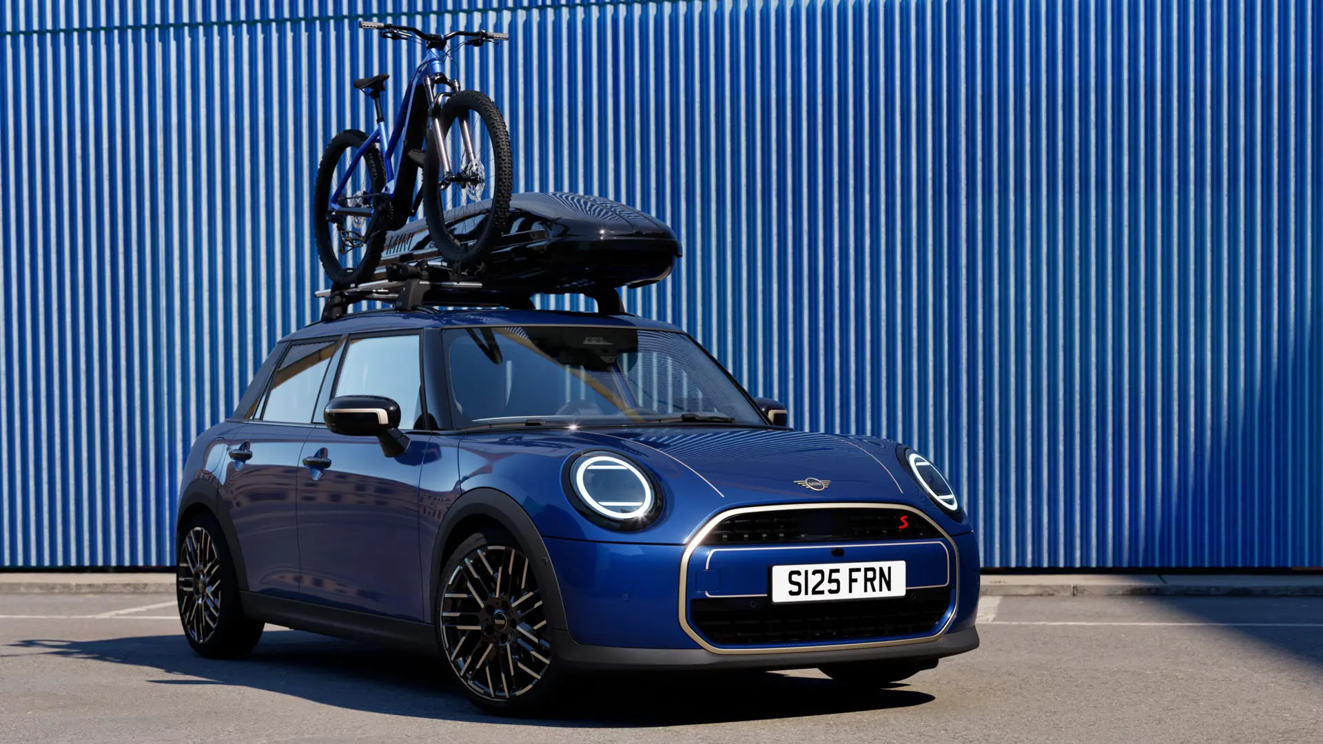 Frontvy av MINI Aceman i Indigo Sunset Blue med MINI takcykelhållare, med en mountainbike, och MINI takbox 320.