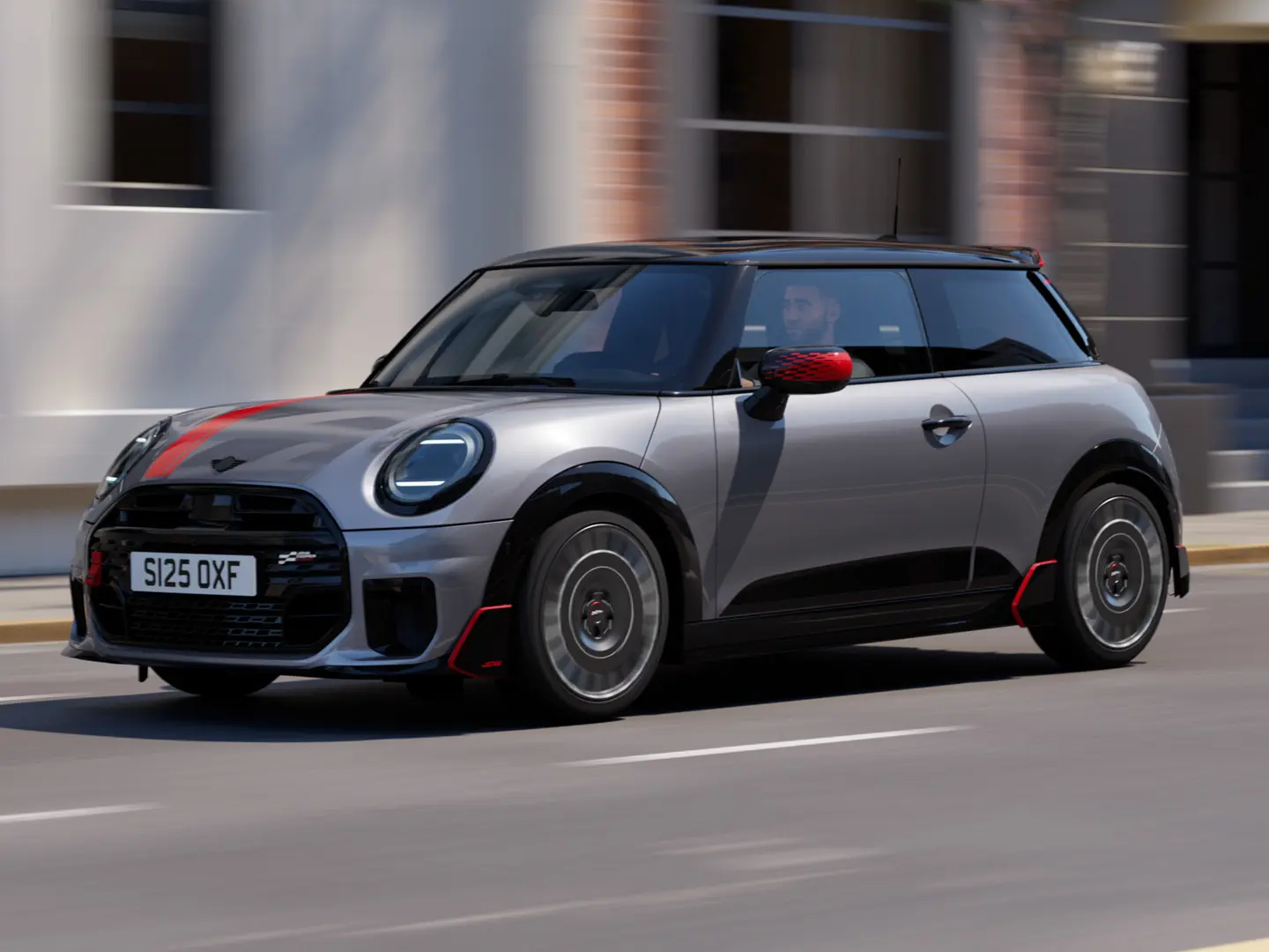 MINI Cooper 3-dörrars i Melting Silver med John Cooper Works backspegelkåpa och motorhuvsdekal i Chili Red, takspoiler och winglet i svart kör längs en gata i stan – vy framifrån från sidan.