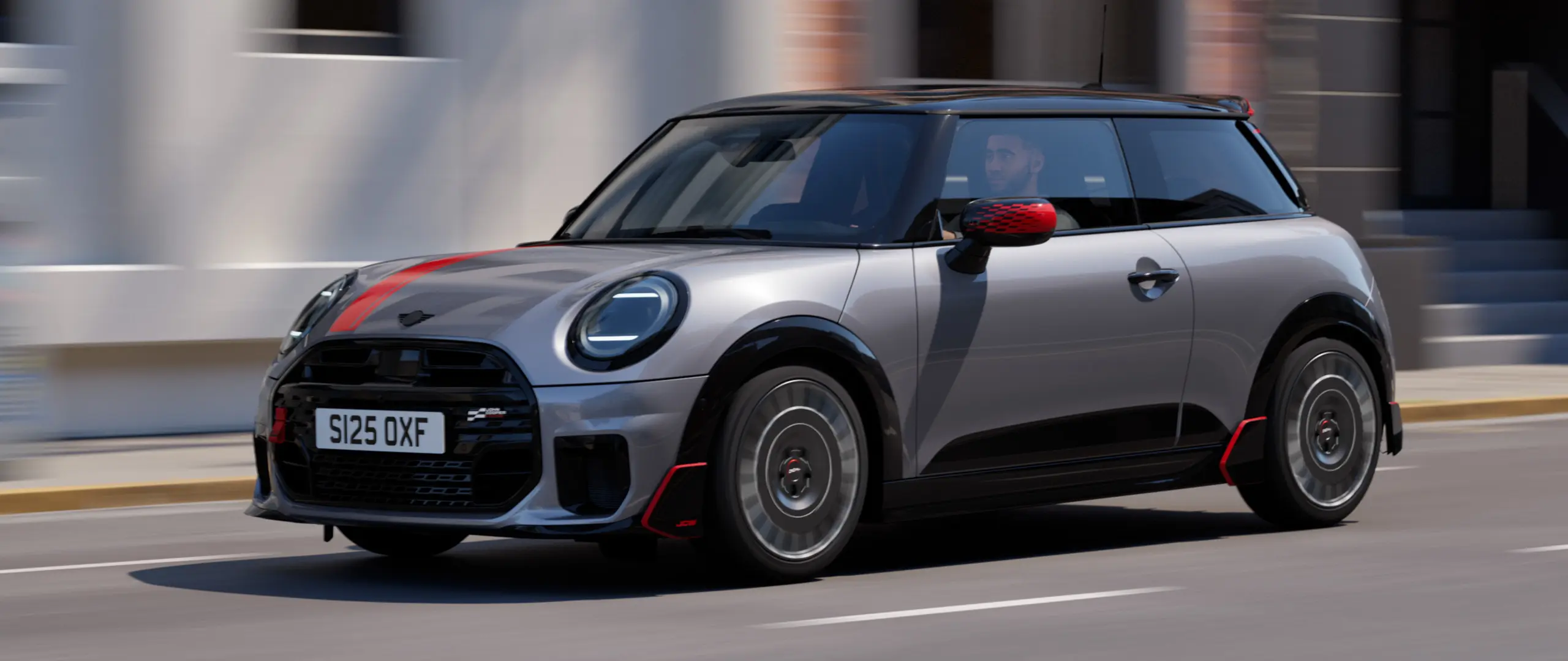 MINI Cooper 3-dörrars i Melting Silver med John Cooper Works backspegelkåpa och motorhuvsdekal i Chili Red, takspoiler och winglet i svart kör längs en gata i stan – vy framifrån från sidan.