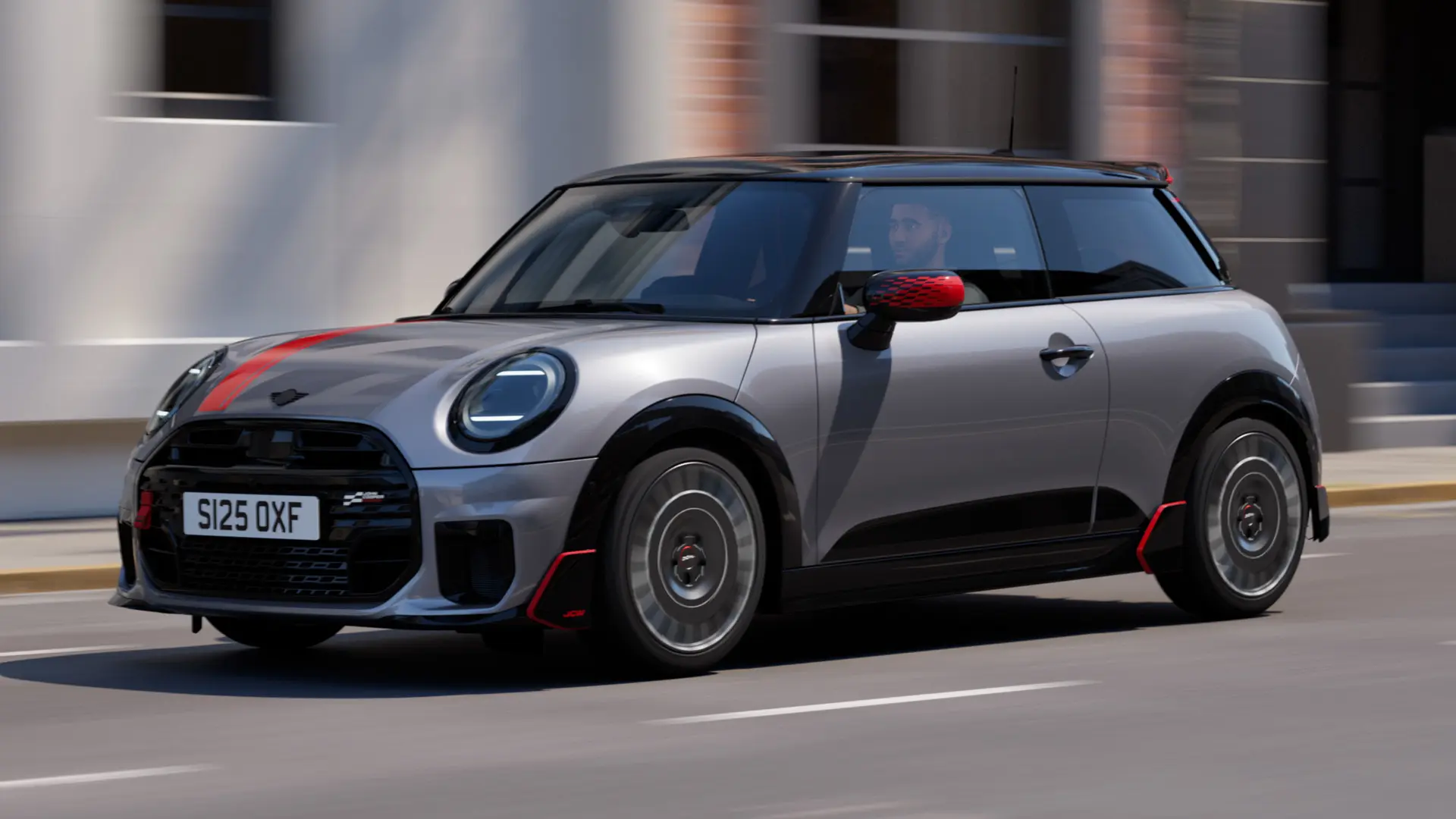MINI Cooper 3-dörrars i Melting Silver med John Cooper Works backspegelkåpa och motorhuvsdekal i Chili Red, takspoiler och winglet i svart kör längs en gata i stan – vy framifrån från sidan.